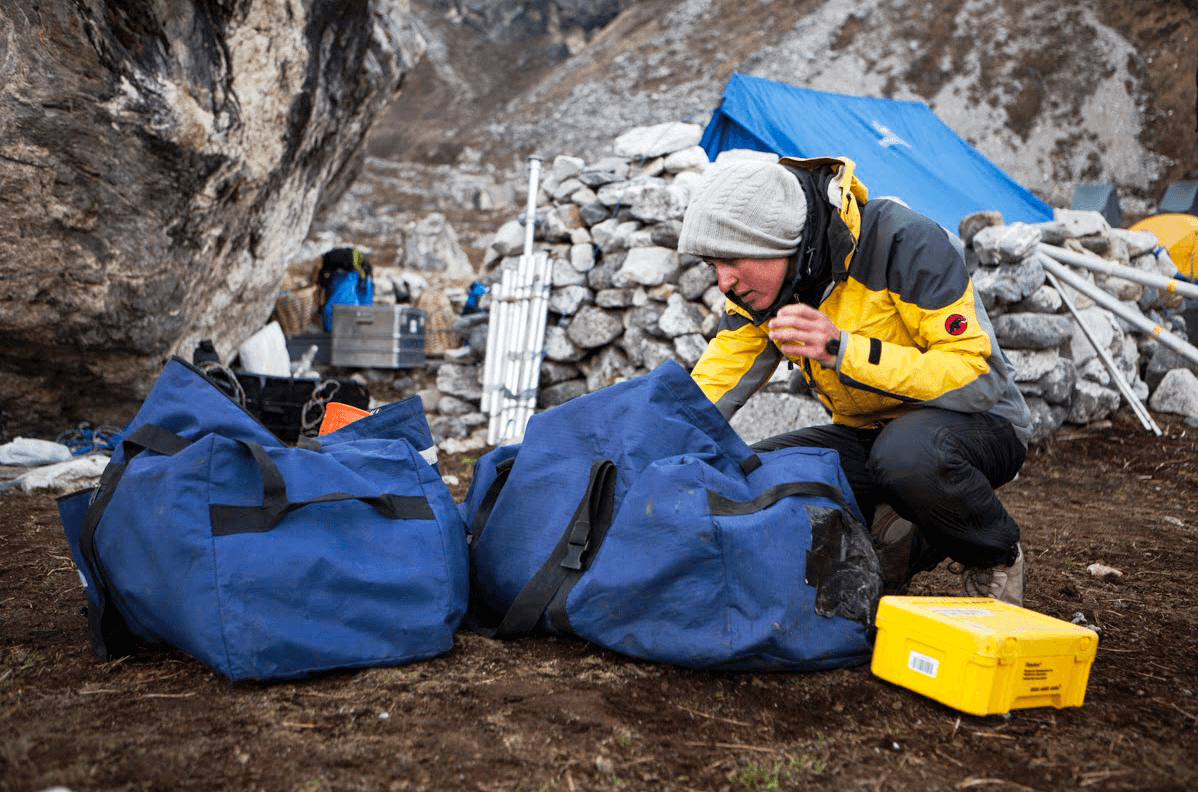 Person in gelber Jacke und Mütze packt blaue Expeditionssäcke in einem Berglager.