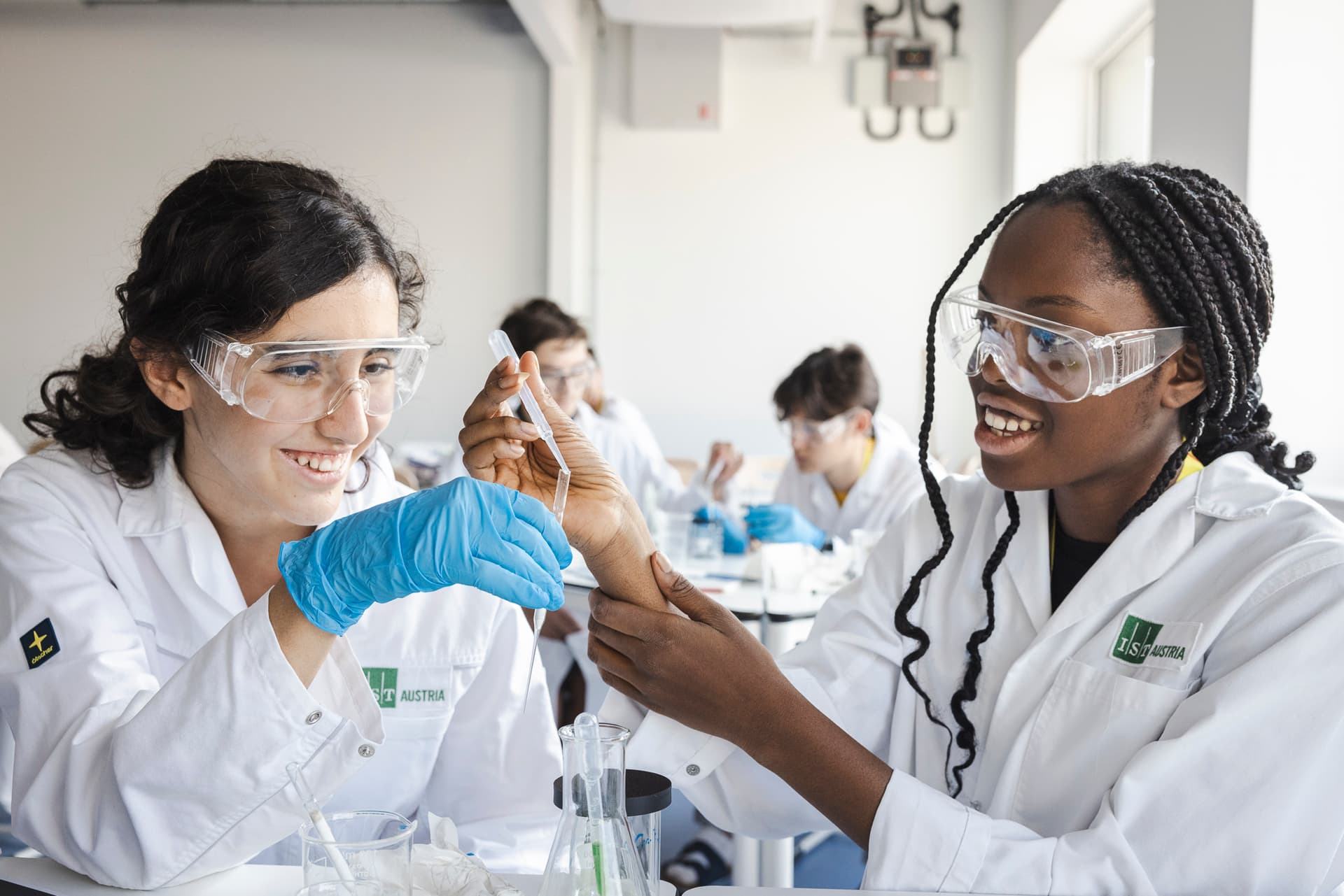 Zwei junge Frauen in Laborkitteln und Schutzbrillen arbeiten gemeinsam an einem wissenschaftlichen Experiment. Eine hält eine Pipette, während die andere ihre Hand unterstützt. Beide lachen und wirken motiviert. Im Hintergrund sitzen weitere junge Menschen in ähnlicher Schutzkleidung an Labortischen.