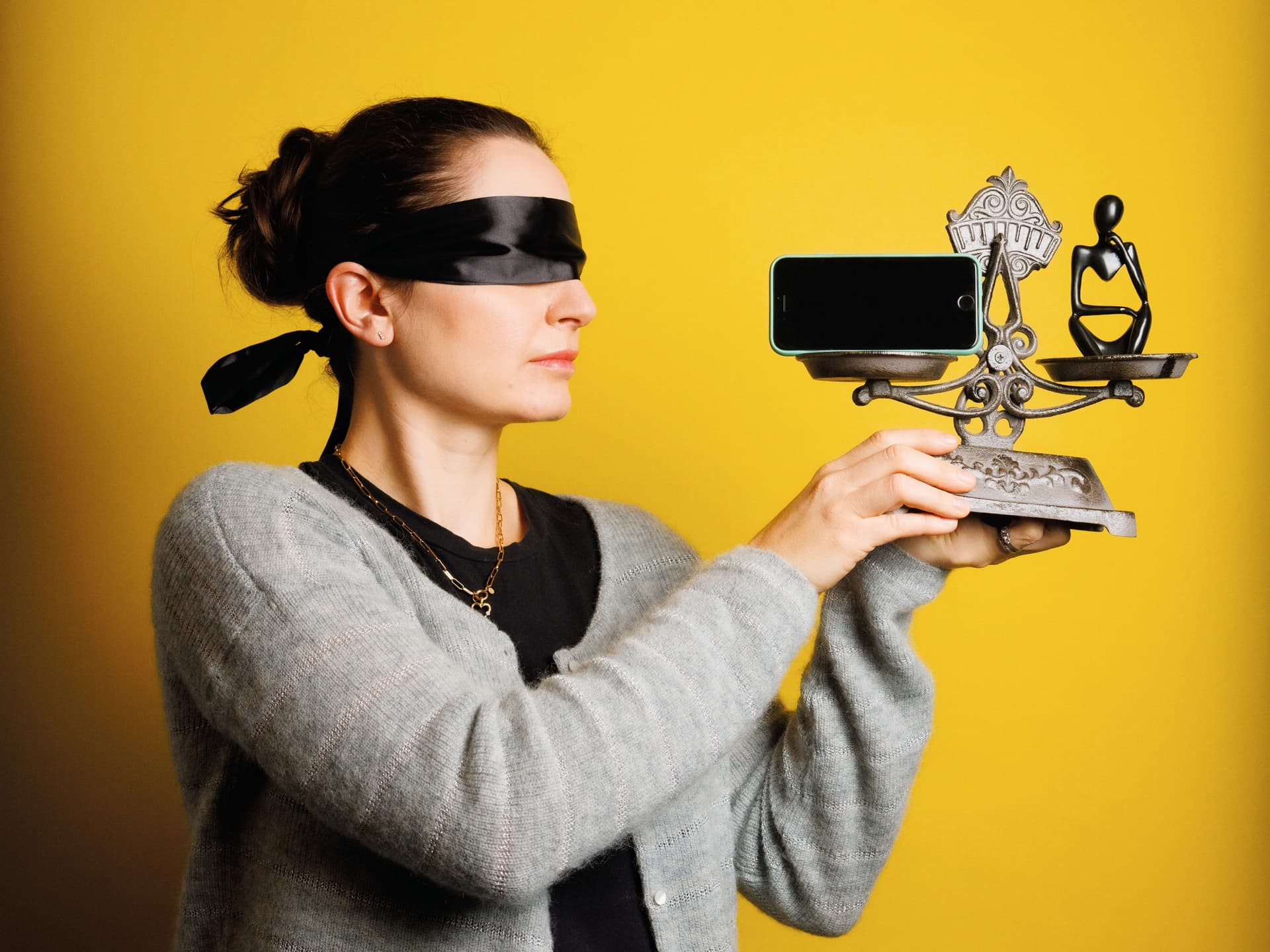 Eine Frau mit von einem schwarzen Band bedeckten Augen hält einen verzierten Ständer mit einem Smartphone vor einem gelben Hintergrund.