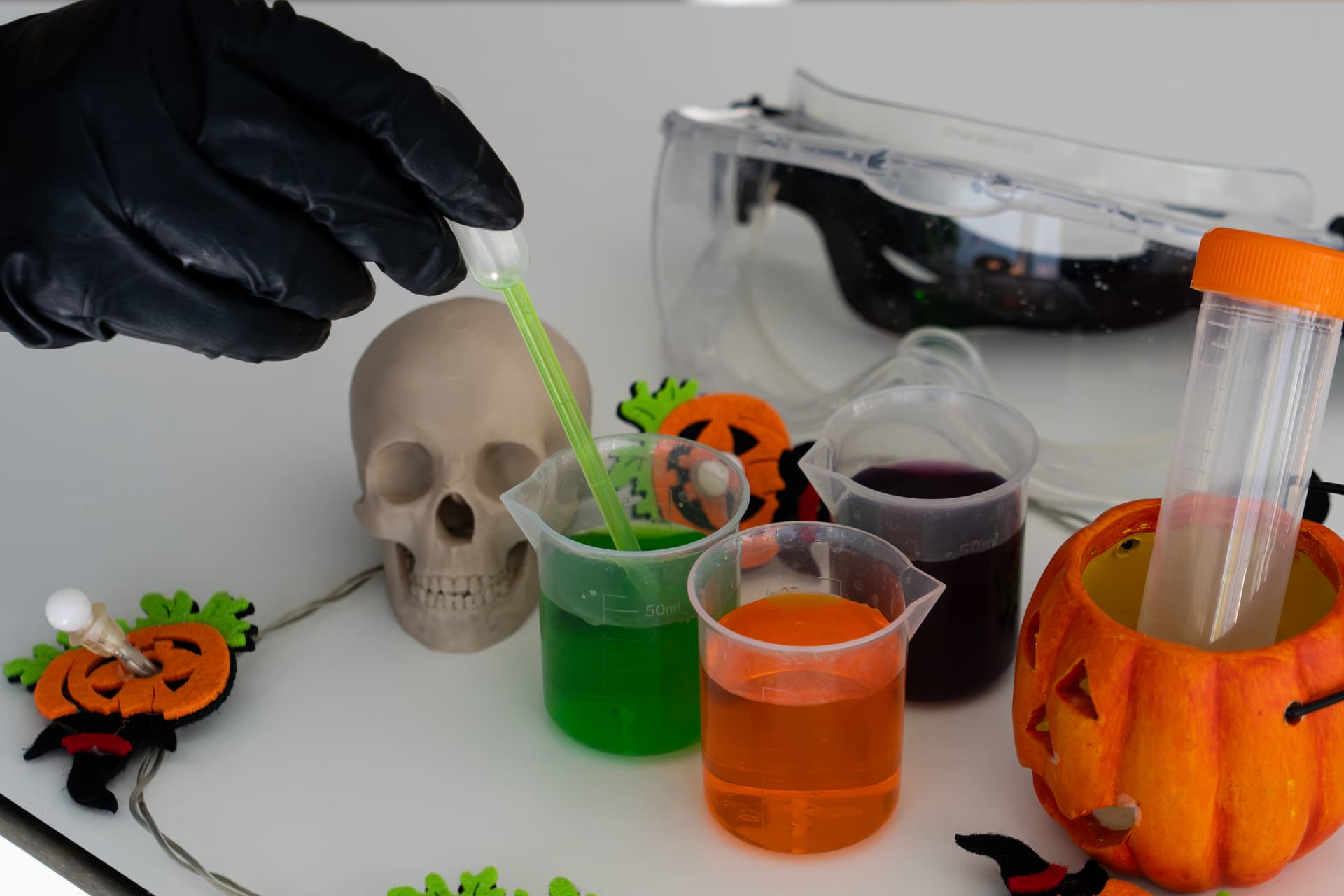 Eine Hand in einem Handschuh bedient eine Pipette mit bunten Flüssigkeiten in Bechern, umgeben von Halloween-Dekorationen, einem Totenkopf und einer Schutzbrille.